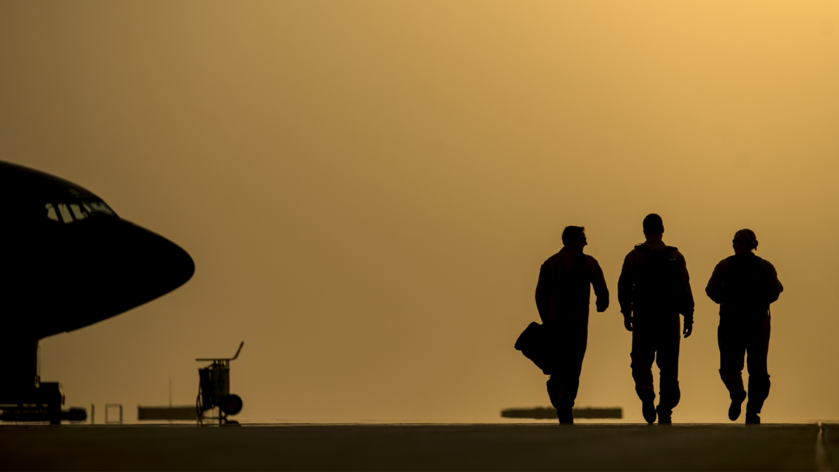 foto con colori sabbia, muso di aeroplano e tre piloti e tecnici che camminano sulla pista visti di schiena